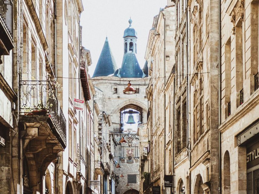 Una estrecha calle europea con edificios de piedra conduce a una caseta de vigilancia histórica con una gran campana visible bajo su arco.