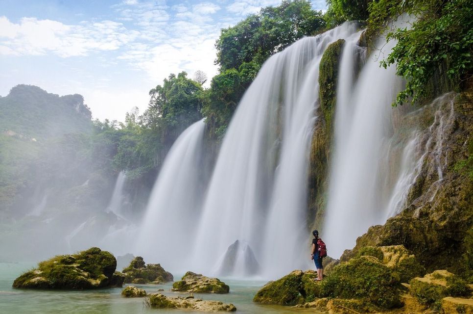 Une personne avec un sac à dos se tient sur des rochers moussus au pied d'une grande cascade, entourée d'un feuillage vert.