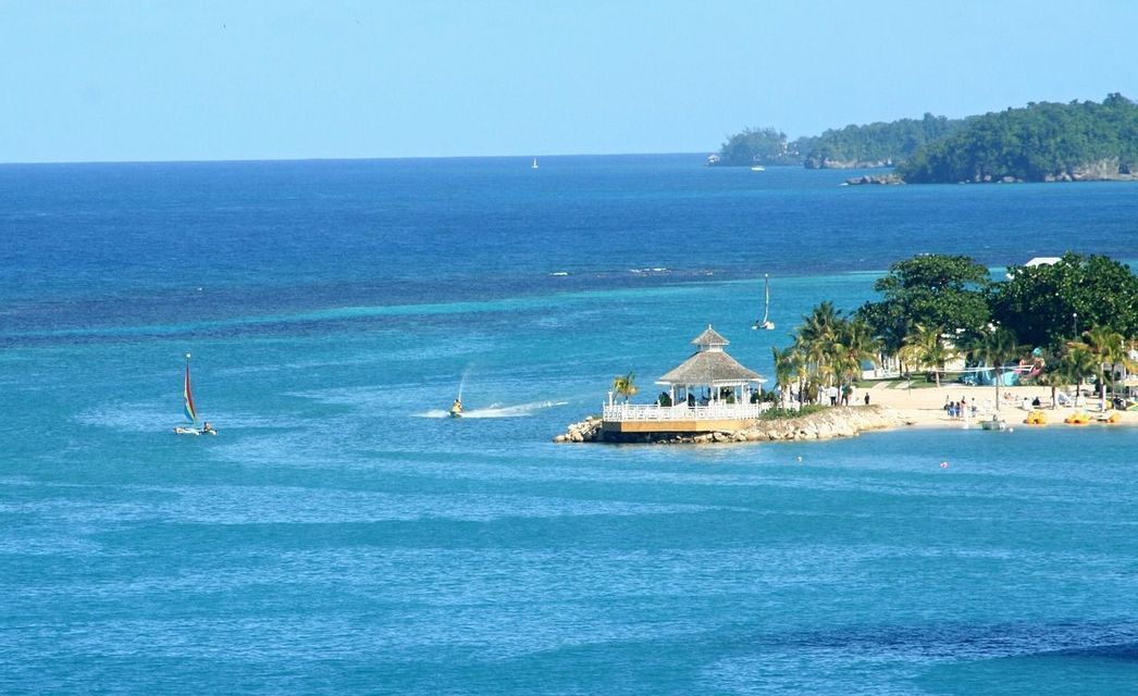 Un kiosque blanc sur une presqu'île domine une plage tropicale avec palmiers et bateaux sur la mer turquoise sous un ciel dégagé.