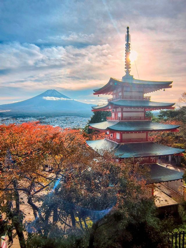 Una pagoda rossa giapponese sorge tra alberi dal fogliame autunnale, affacciata su una città con un grande vulcano sullo sfondo all'alba.