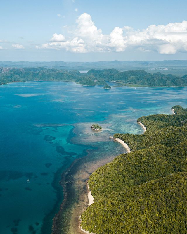 Vista aerea di una costa tropicale con lussureggianti colline verdi che si affacciano su un oceano turchese e isole in lontananza.