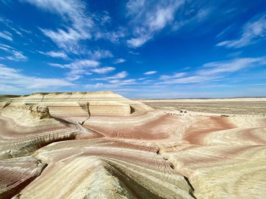 Formazioni rocciose colorate e stratificate si estendono in un deserto sotto un cielo azzurro brillante con nuvole e due veicoli in lontananza.