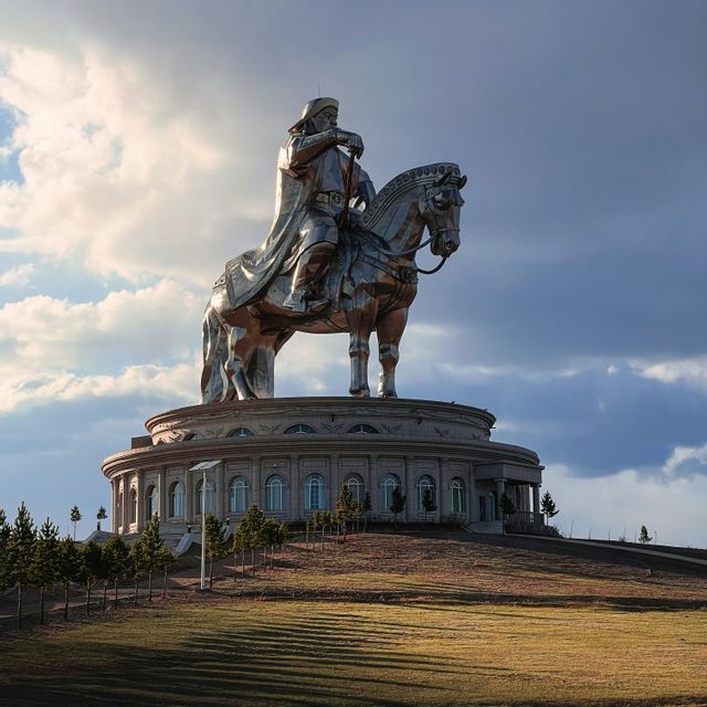Una gigantesca statua equestre in argento di Gengis Khan si erge in cima a un grande edificio circolare su una collina erbosa sotto un cielo parzialmente nuvoloso.