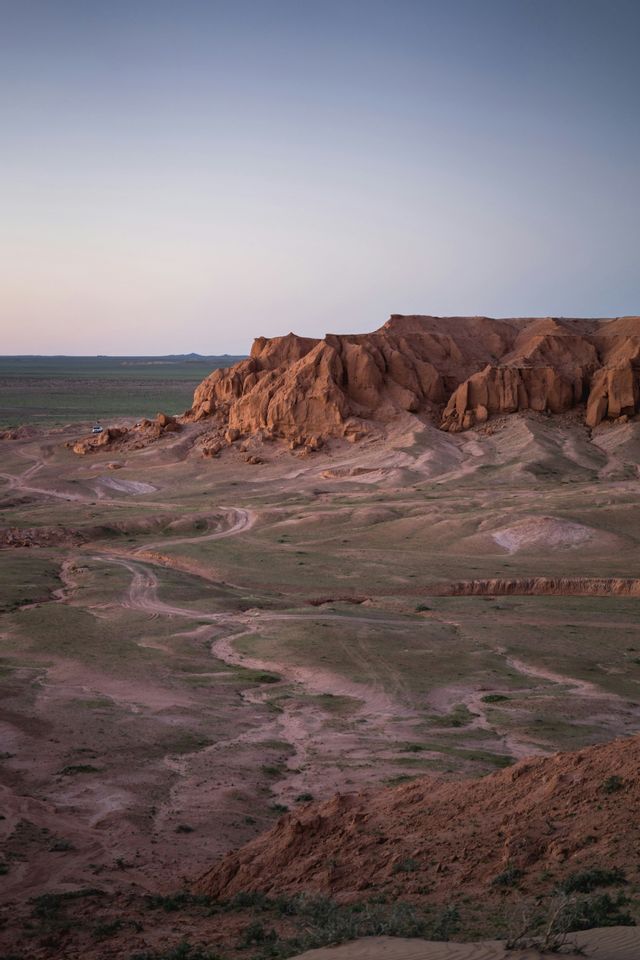 Un vasto paesaggio desertico con grandi formazioni rocciose rosse e tortuosi sentieri sterrati sotto un cielo pallido al tramonto.