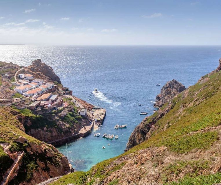 Vista aerea di un borgo costiero con case bianche incastonate in una cala rocciosa e verde. Le barche sono ormeggiate nella baia turchese accanto a un pontile.