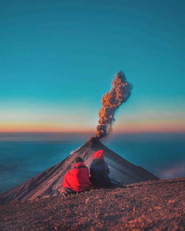 Two people sit on a mountainside watching an erupting volcano send a plume of smoke into the sunrise sky.