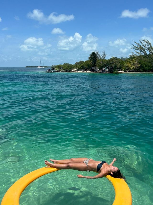 A woman in a bikini floating on her back on a yellow object in clear turquoise water, with a tropical island in the background.
