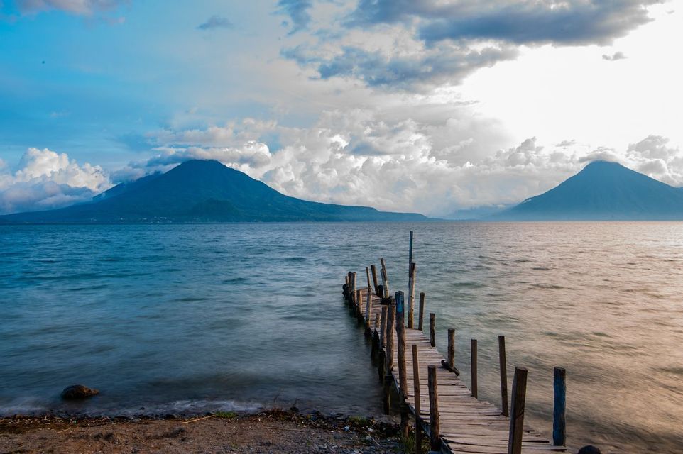 A rustic wooden pier extends into a large lake with two mountains in the background under a cloudy sky.