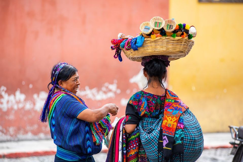 Two women in colorful traditional clothing on a street, one balances a woven basket full of goods on her head.