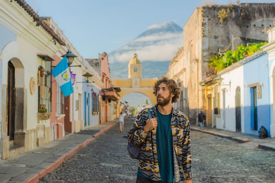 A man with a backpack stands on a cobblestone street lined with colorful buildings, with a yellow arch and a large volcano in the background.