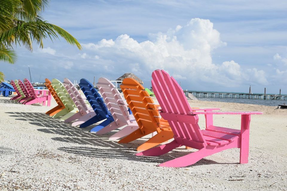 A row of colorful wooden Adirondack chairs sit on a sandy beach facing the ocean under a partly cloudy sky.
