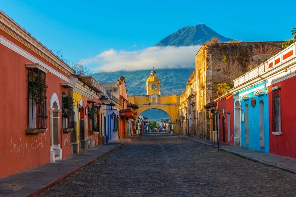 A cobblestone street lined with colorful colonial buildings leads to a yellow archway with a clock, with a volcano in the background under a blue sky.