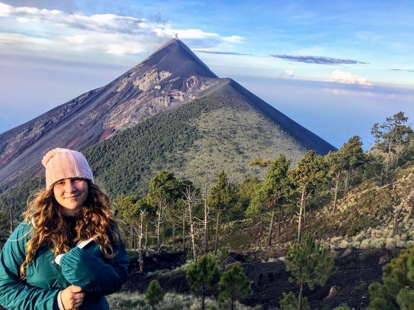 A woman in a pink beanie and green jacket smiles on a mountainside, with an active volcano erupting smoke in the background.
