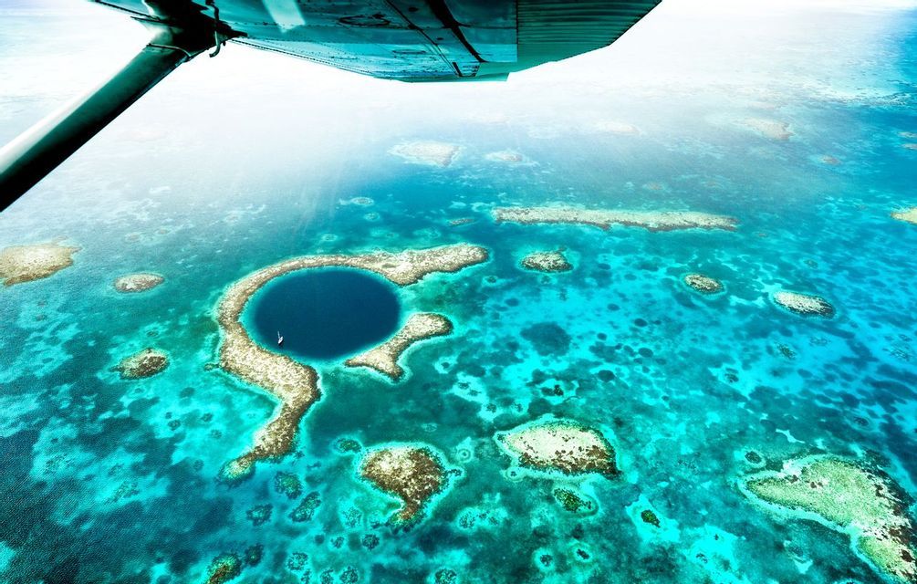 An aerial view from under an airplane wing of a large blue hole surrounded by coral reefs in a turquoise ocean.