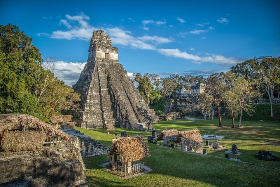 An ancient stone pyramid and thatched-roof structures on a grassy clearing in the jungle under a partly cloudy blue sky.