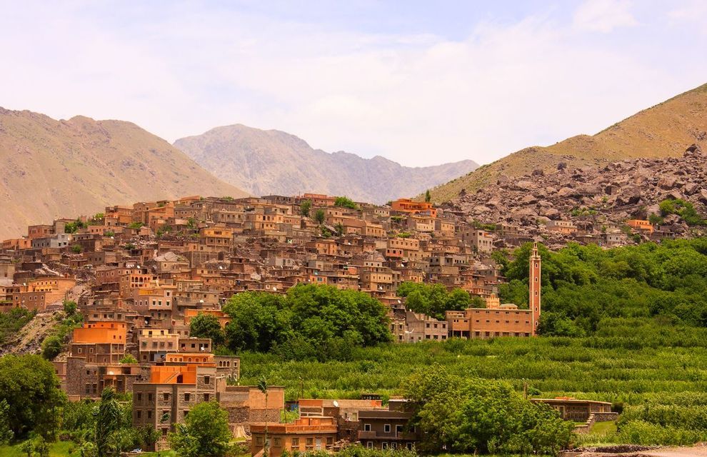 An earthen-colored village built on a lush, green hillside at the base of rocky mountains.
