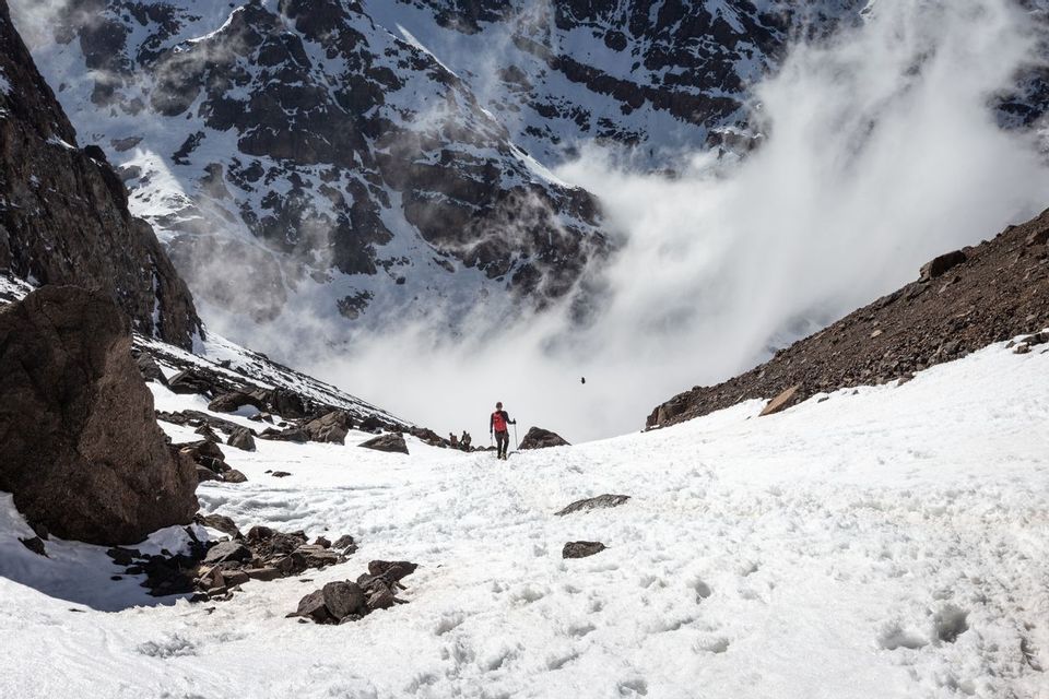 A WeRoad group trip hikes up a snowy trail in a rocky mountain valley under a partly cloudy sky.