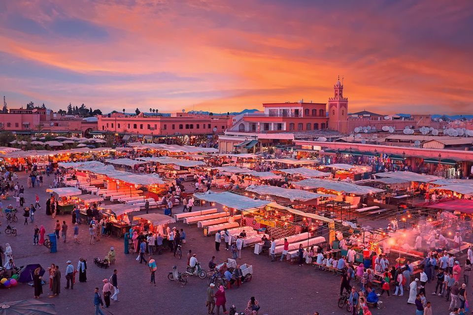 An elevated view of a bustling outdoor market square filled with illuminated food stalls and crowds of people at sunset.