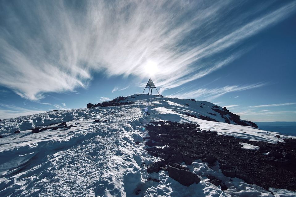 A snowy mountain path leads to a triangular summit marker, with the sun shining brightly behind it under a cloudy blue sky.