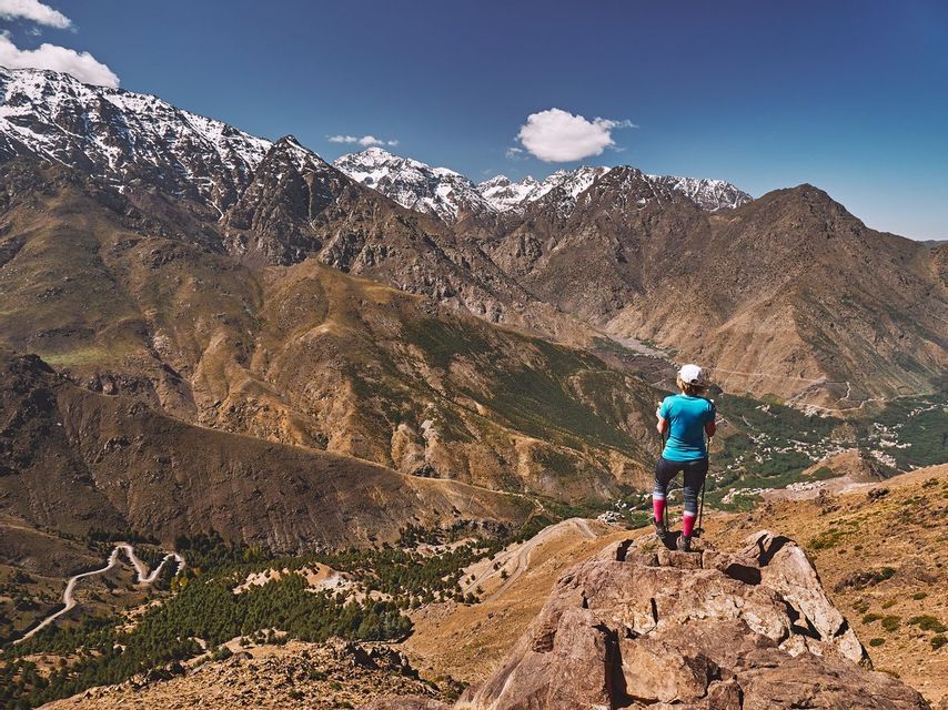A hiker stands on a rocky peak, looking out at a panoramic view of snow-capped mountains and a valley with a winding road.