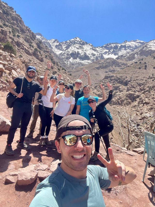 A WeRoad group trip takes a selfie while hiking on a rocky mountain trail, with snow-capped peaks behind them.