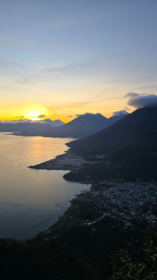 Vista elevada de un pueblo a orillas del lago rodeado de montañas al amanecer, con el resplandor del sol en el horizonte.