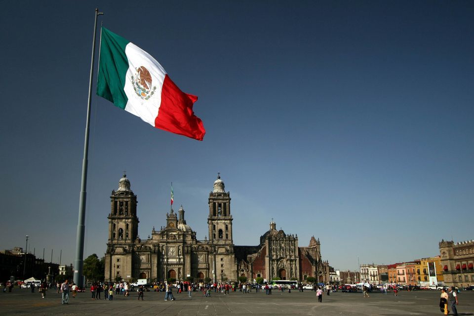 The Mexican flag waves on a tall flagpole in a busy public square, with a large, historic cathedral in the background.