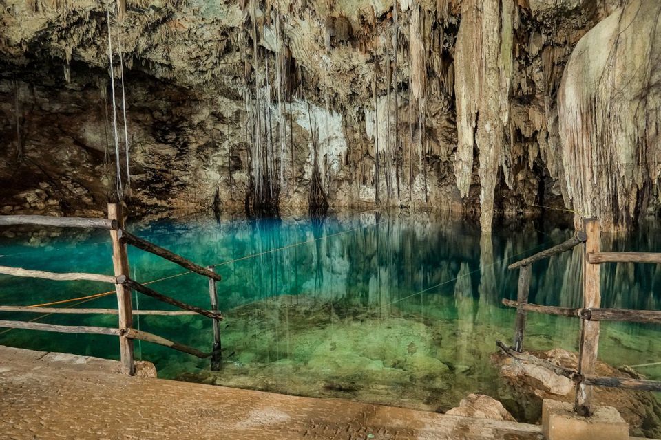 A natural cenote inside a cave with clear, turquoise water reflecting the stalactites hanging from the ceiling. A wooden fence is in the foreground.