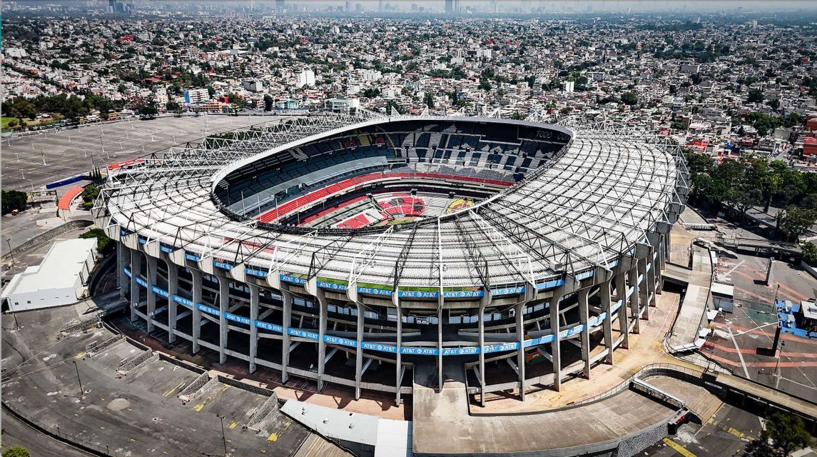 An aerial view of a large, empty football stadium with a complex roof structure, surrounded by a vast, sprawling cityscape.