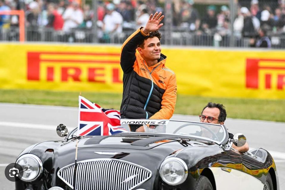 A man in an orange and black jacket waves to a crowd while standing in a classic black convertible car with a British flag.