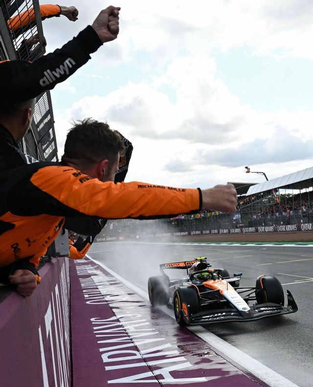 A McLaren Formula 1 car races on a wet track, spraying water as team members in orange cheer from over a barrier.