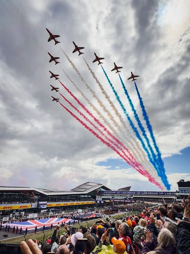 Nine jets fly in formation, leaving trails of red, white, and blue smoke over a crowded grandstand during an airshow.