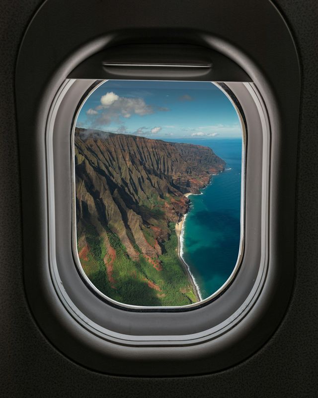 View from an airplane window overlooking a rugged coastline with steep green cliffs dropping into a turquoise ocean.
