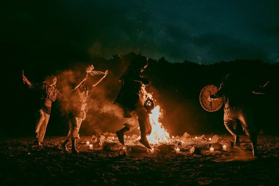 A group of people in traditional clothing performs a ritual dance around a large bonfire on the sand at night.