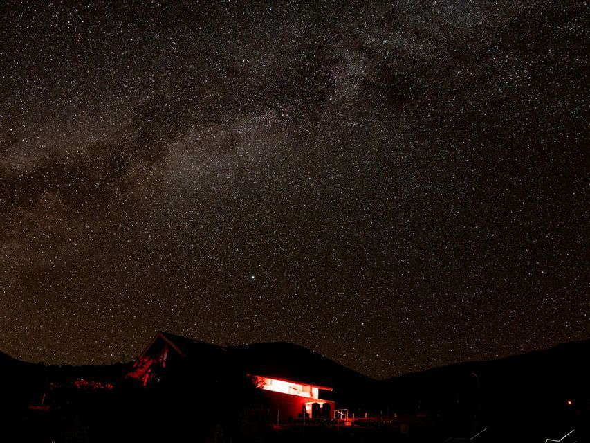 A vast, starry night sky with the Milky Way galaxy visible above a silhouetted house illuminated by red light.