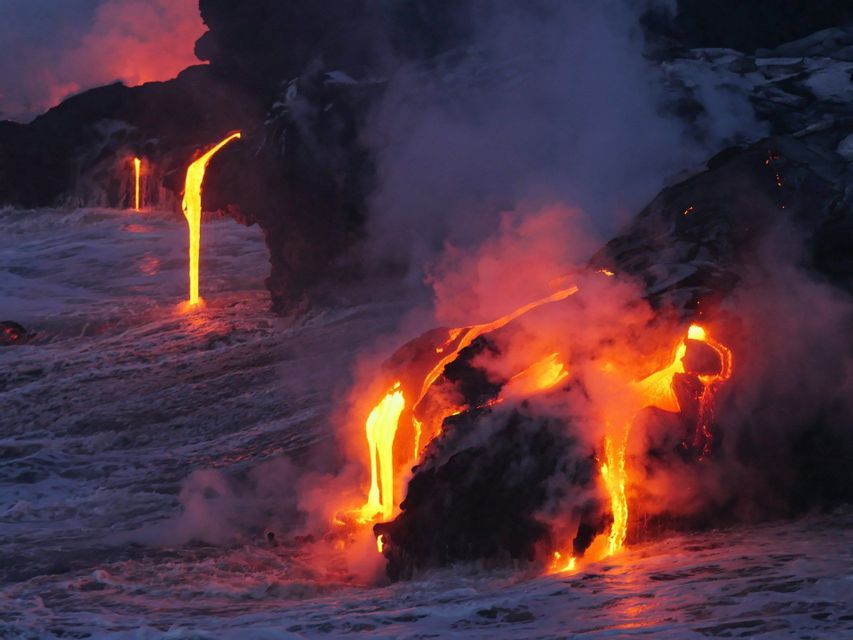 Molten lava flows from dark volcanic rocks into the churning ocean, creating clouds of steam at dusk.