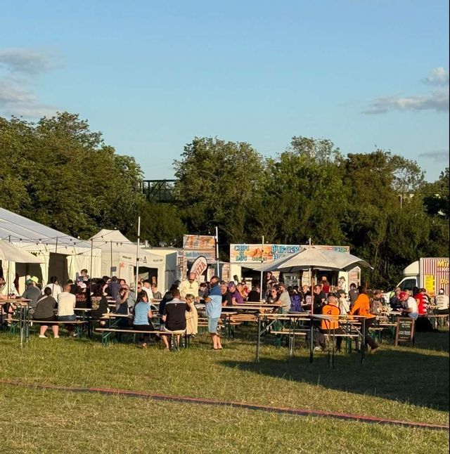 A crowd of people eating and socializing at long wooden tables during an outdoor food festival on a grassy field.