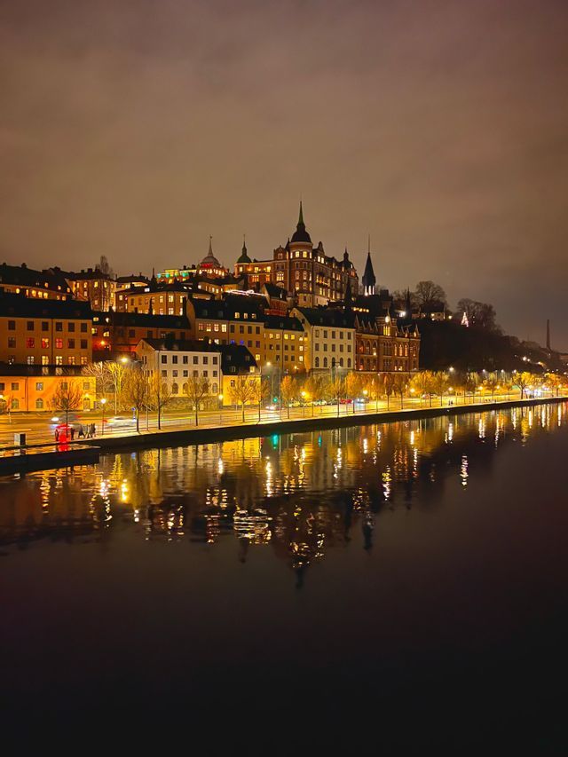Skyline notturno di una città sul lungomare, con le luci calde di edifici e strade che si riflettono sull'acqua scura.