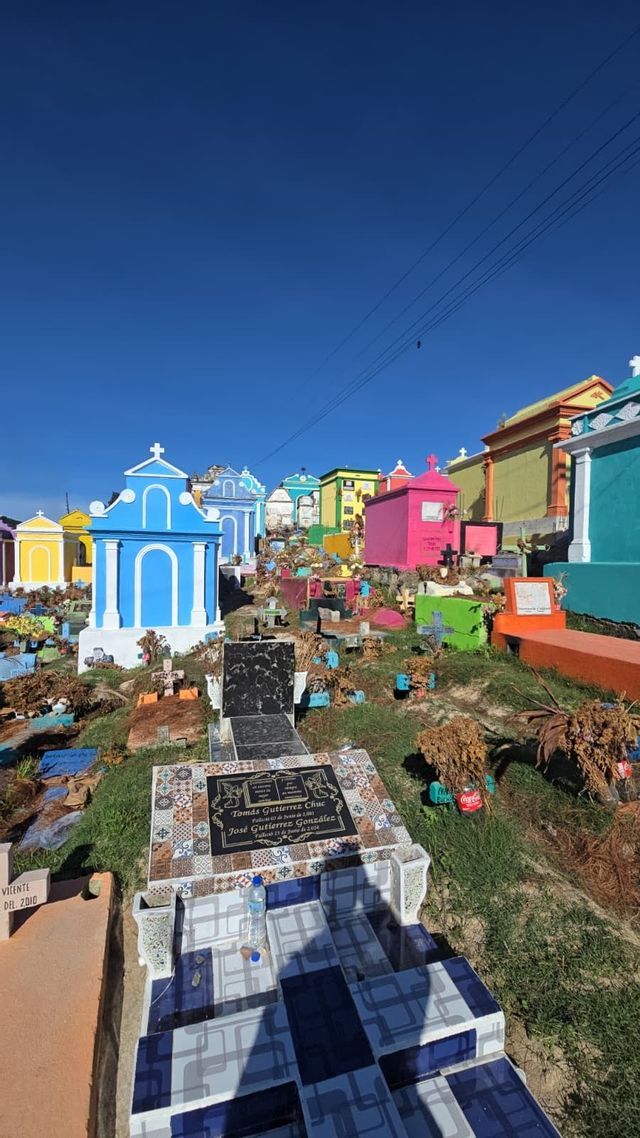 Un vibrante cementerio en una ladera con mausoleos de colores brillantes, en azul, rosa y amarillo, bajo un cielo azul claro.