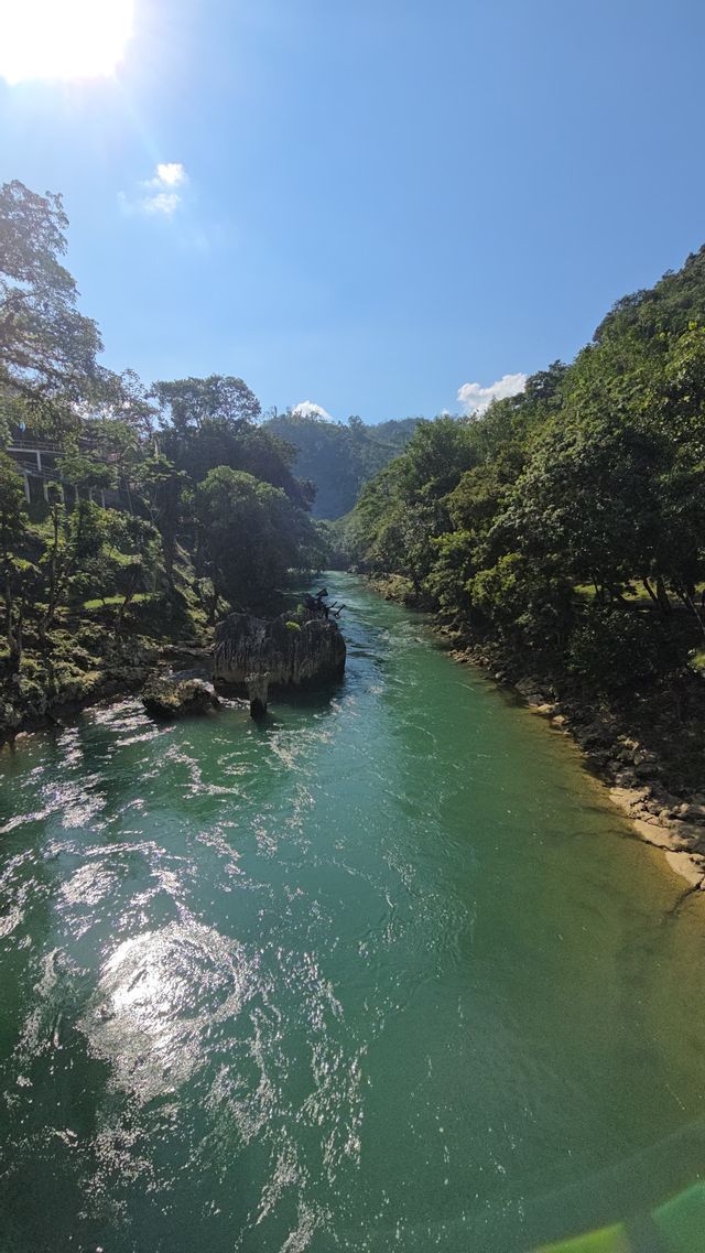 Un río turquesa fluye a través de un valle con exuberantes colinas verdes a ambos lados, bajo un cielo azul soleado.
