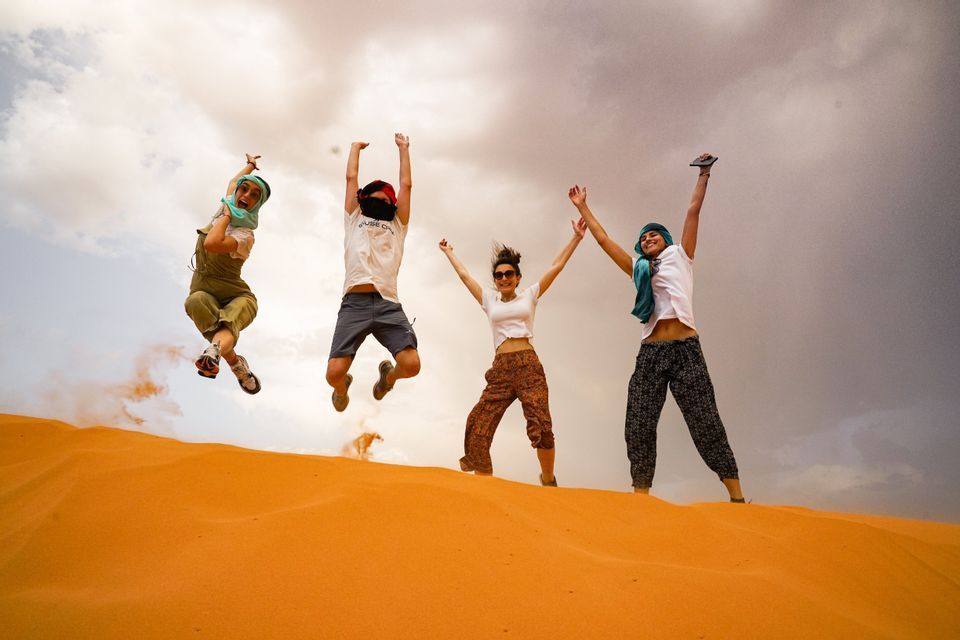 A WeRoad group trip of four people jumping together with their arms up on top of an orange sand dune.