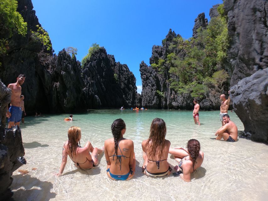 A WeRoad group trip relaxing in the clear shallow water of a lagoon surrounded by tall, dark rock formations under a sunny sky.