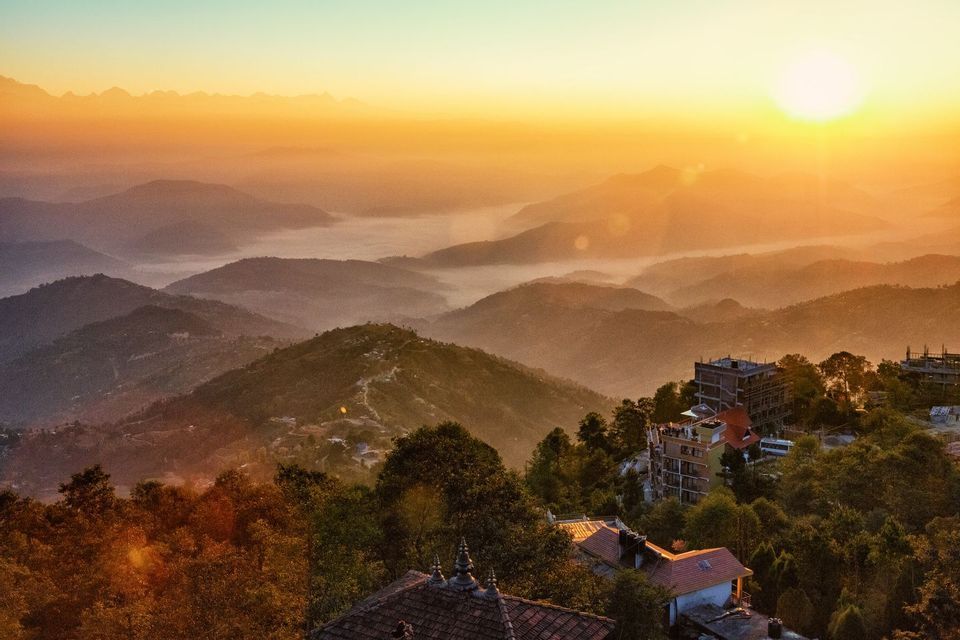 Un'alba dorata illumina montagne e valli nebbiose e stratificate, immerse nella nebbia, con edifici annidati tra gli alberi in primo piano.