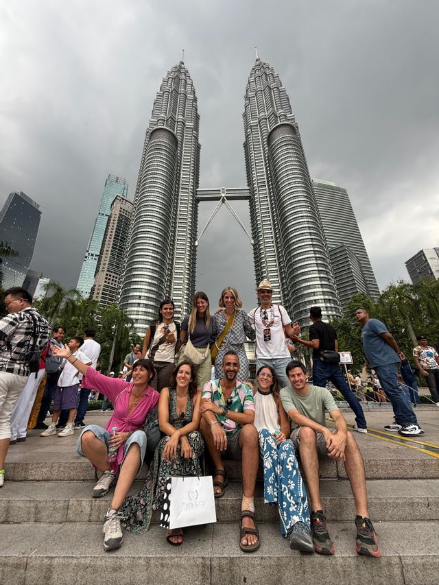 Eine WeRoad Gruppenreise posiert für ein Foto auf Stufen vor hohen Zwillingswolkenkratzern unter einem bewölkten Himmel.