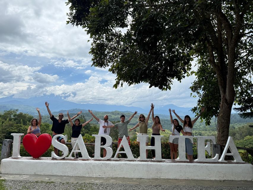 Un viaje en grupo de WeRoad posando con los brazos levantados detrás de un gran cartel de 'I Love Sabah Tea' con un paisaje montañoso de fondo.