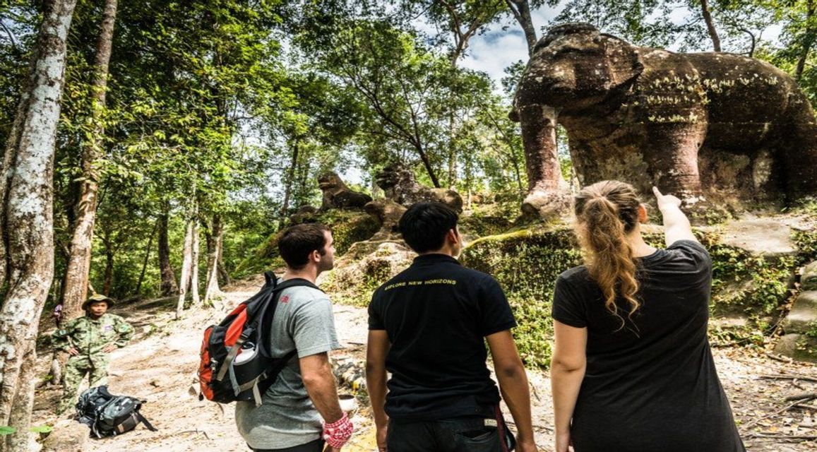Una mujer de un viaje en grupo de WeRoad señala una gran estatua de elefante de piedra cubierta de musgo mientras dos hombres observan en un bosque.