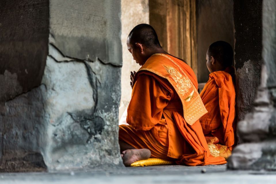 Due monaci in abiti arancioni sono seduti e pregano sul pavimento all'interno di un tempio di pietra consumato dal tempo.