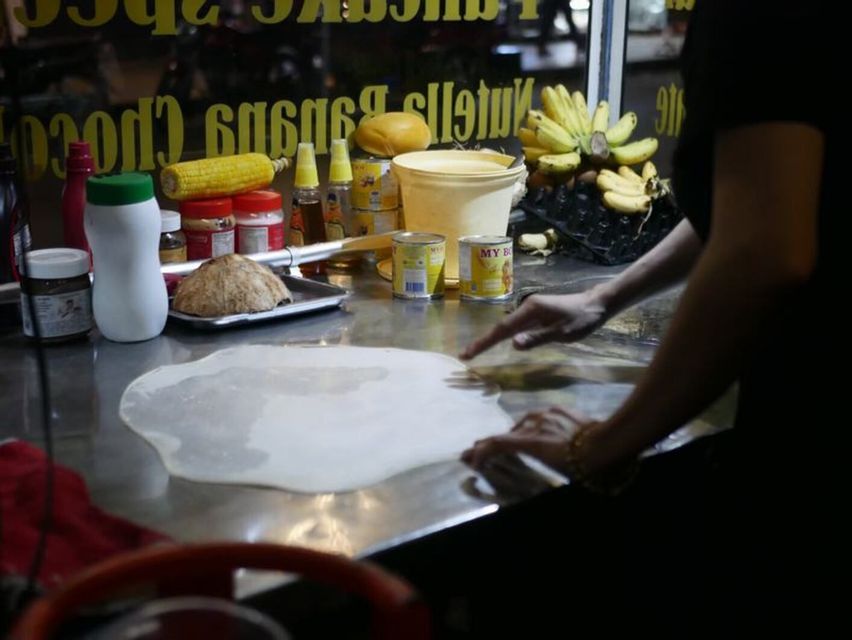 Una persona estira una fina lámina circular de masa sobre un mostrador de metal en un puesto de comida nocturno, rodeada de ingredientes como plátanos y maíz.