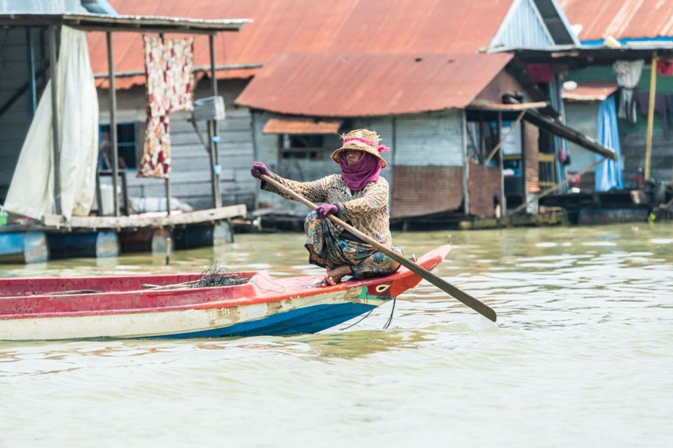 Una persona con sombrero de paja y mascarilla rema en una barca pequeña en un pueblo flotante con casas sobre pilotes.
