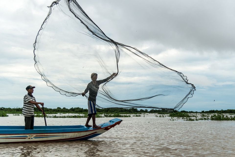 Due pescatori in una piccola barca su un fiume, uno in piedi a prua che lancia una grande rete da pesca.
